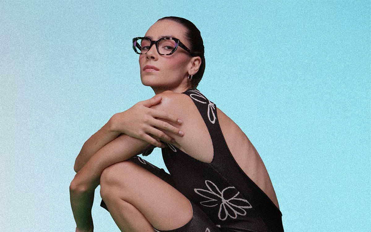 A woman crouching down in front of a blue background wearing Onirico glasses with a bold shaped frame.